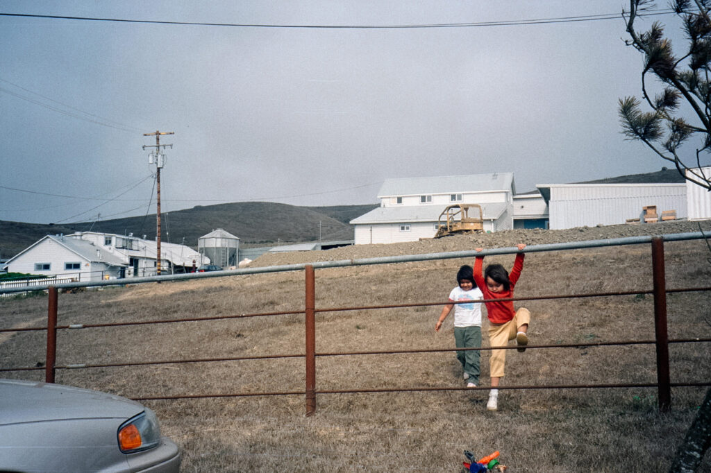 Two children playing in a fence in a field with white barn-style structures in the background.